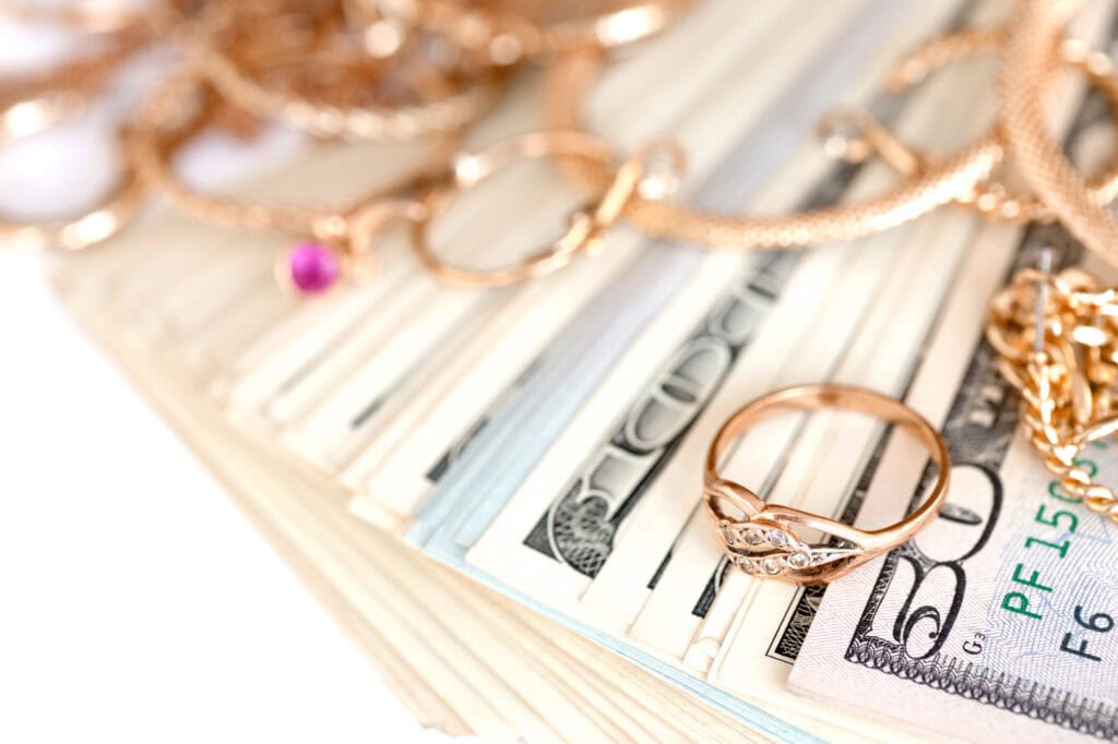 Many gold jewelry rings, earrings and necklaces with big amount of US dollar bills on white background.  How To Sell Gold (Step-By-Step Guide)