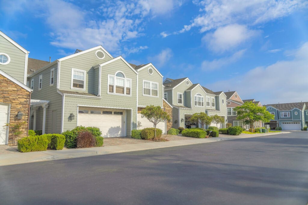 Duplex houses with green wood vinyl sidings at Carlsbad, San Diego, California. There are white garage doors with concrete driveway near the plants at the front. how to make $30,000 a month with real estate